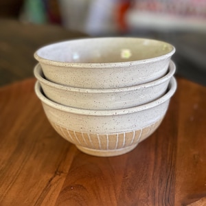 May include: A stack of three off-white ceramic bowls with speckled detailing. The bottom portion of the bowls features a textured, vertical striped design. The bowls are stacked on a wooden serving tray, creating a rustic aesthetic.