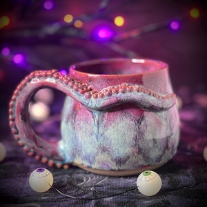 Peut inclure: Une tasse en céramique avec un vernis brillant rose et bleu, dotée d'une anse sculptée en forme de tentacule de pieuvre couverte de petites ventouses. Deux petits objets sphériques blancs avec des iris peints en violet et vert sont posés devant.