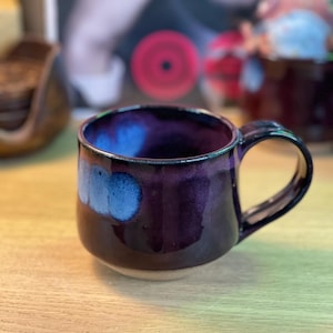 May include: A handmade ceramic mug with a dark purple glaze and blue accents. The mug has a rounded shape with a black handle and a light-colored base. The interior is a vibrant blue.