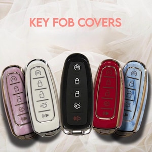 May include: A collection of key fob covers in various colors, including pink, white, black, red, and blue. The covers have a sleek design with metallic accents and feature the text "KEY FOB COVERS". Each cover displays the key functions.