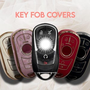 May include: A collection of key fob covers in various colors, including cream, pink, red, and black. The covers have gold or black trim and feature different button designs. The text "KEY FOB COVERS" is displayed in pink.