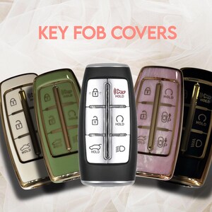 May include: A collection of key fob covers in various colors, including white, green, pink, and black. Each cover has a metallic gold trim and features the text "HOLD" and other key symbols. The text "KEY FOB COVERS" is displayed in pink.