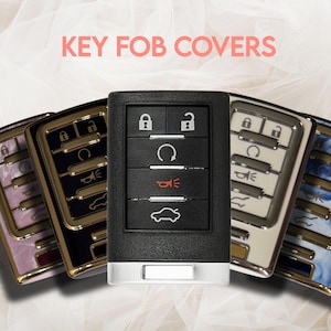 May include: A collection of key fob covers in various colors and designs. The central black key fob is surrounded by covers with gold trim and patterns in pink, black, white, and blue. The text "KEY FOB COVERS" is displayed in pink.