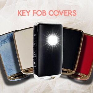 May include: A collection of key fob covers in various colors, including blue marble, cream, black, and red. Each cover has a gold-colored trim and a bright light emanating from the center. The text "KEY FOB COVERS" is displayed at the top.