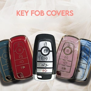 May include: Image of key fob covers in various colors, including red, blue, and pink, with gold-colored trim. The text "KEY FOB COVERS" is displayed in pink at the top. The key fobs have a metallic finish and feature button details.