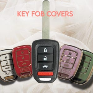 May include: A collection of key fobs in various colors, including red, white, pink, and green. The key fobs have a metallic trim and feature button designs for locking, unlocking, and other functions. The text "KEY FOB COVERS" is displayed in pink.