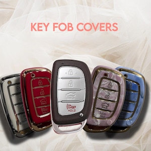 May include: A collection of key fob covers in various colors, including silver, red, brown, pearlescent, and blue. The key fobs have a metallic finish and feature buttons for locking, unlocking, and other functions. The text "KEY FOB COVERS" is displayed in pink.