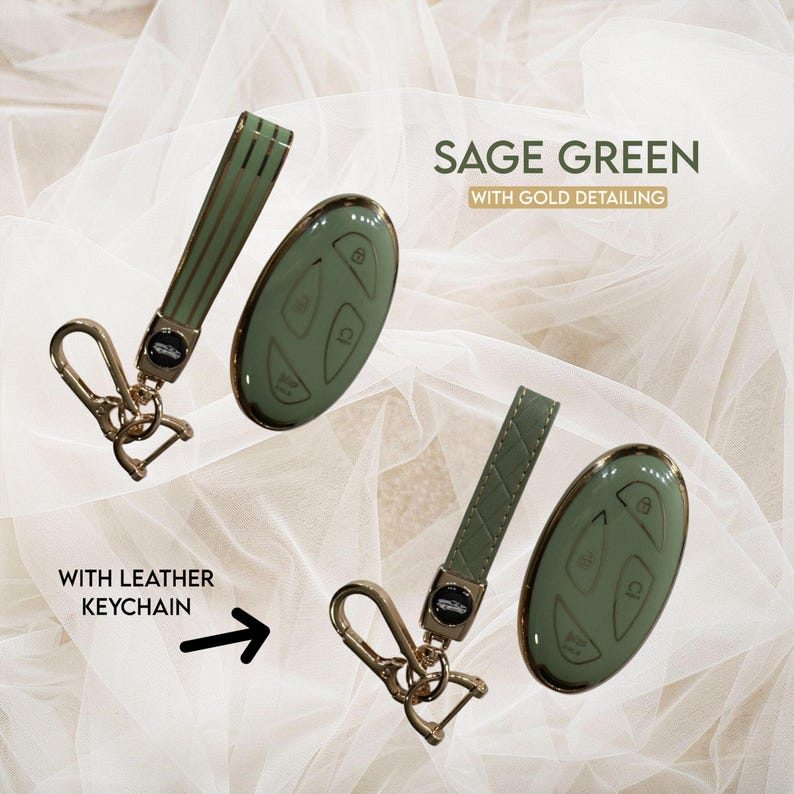May include: Sage green car key fob with gold detailing and a matching leather keychain. The key fob has gold accents and a smooth finish. The keychain has a gold clasp.