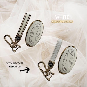 May include: White car key fob with gold detailing and a matching leather keychain. The key fob has buttons for lock, unlock, and hold. The keychain is white with a gold clasp. The text in the image says "White with gold detailing" and "With leather keychain".