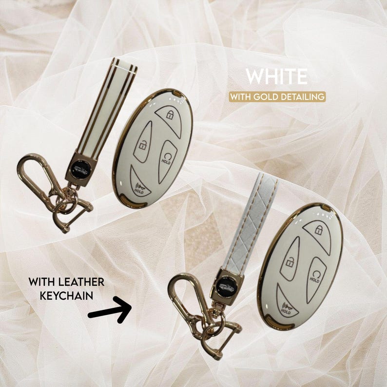 May include: White car key fob with gold detailing and a matching leather keychain. The key fob has buttons for lock, unlock, and hold. The keychain is white with a gold clasp. The text in the image says "White with gold detailing" and "With leather keychain".