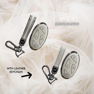 May include: White car key fob with silver detailing and a matching leather keychain. The key fob has buttons for locking, unlocking, and other functions. The keychain has a silver clasp.