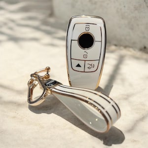 May include: A white car key fob and matching key chain with gold-coloured accents. The key fob has a gold trim and black buttons. The key chain has a white strap with gold stripes and a gold clasp.