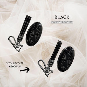 May include: A black car key fob cover with silver detailing and a matching black leather keychain. The set includes a metallic carabiner clip and a key ring. The text "BLACK WITH SILVER DETAILING" and "WITH LEATHER KEYCHAIN" is visible.