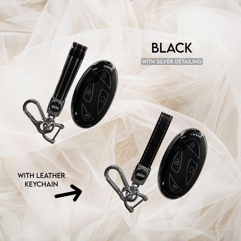 May include: A black car key fob cover with silver detailing and a matching black leather keychain. The set includes a metallic carabiner clip and a key ring. The text "BLACK WITH SILVER DETAILING" and "WITH LEATHER KEYCHAIN" is visible.
