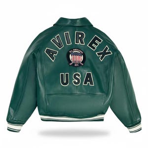 Könnte beinhalten: Grüne Lederjacke mit weißen und schwarzen Akzenten. Auf dem Rücken steht das Wort "AVIREX" in einem Bogen, darunter "USA" und ein Aufnäher. Die Jacke hat einen Kragen, gerippte Bündchen und einen weißen Streifen am Saum.