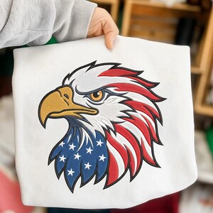 Op de afbeelding: Witte sweatshirt met een geborduurd adelaarskopontwerp. De adelaarskop is gedetailleerd met rode en witte strepen, blauwe sterren en een gele snavel. Het ontwerp is een patriottisch symbool.