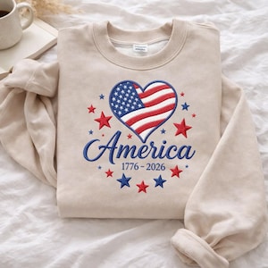 May include: A beige sweatshirt with a patriotic design. The design features a heart-shaped American flag, surrounded by red and blue stars, with the word "America" and the years "1776-2026" embroidered in blue.
