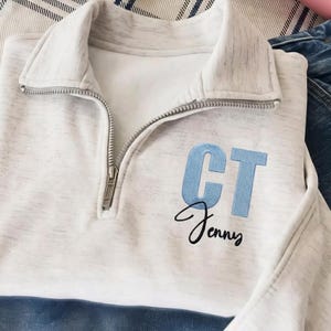 May include: A light gray heathered quarter-zip pullover featuring a silver metal zipper. The garment is embroidered with the light blue block letters CT and the black cursive name Jenny positioned directly below the monogram on the chest.