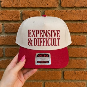 May include: A cream-colored five-panel baseball cap with a flat red brim. The front features the embroidered text "EXPENSIVE & DIFFICULT" in red. A silver and black sticker on the brim reads "OTTO CLASSIC 5 PANEL MID PROFILE MAXIMUM COMFORT".