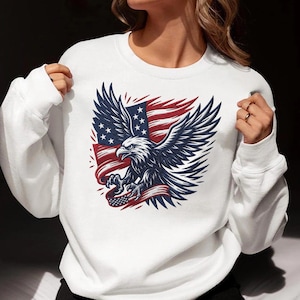 Könnte beinhalten: Weißes Sweatshirt mit einer Grafik eines amerikanischen Weißkopfseeadlers mit ausgebreiteten Flügeln, der ein Banner mit dem Design der amerikanischen Flagge hält. Der Adler ist in Blau und Weiß gehalten, vor einem Hintergrund aus rot-weiß gestreiften Flaggen.