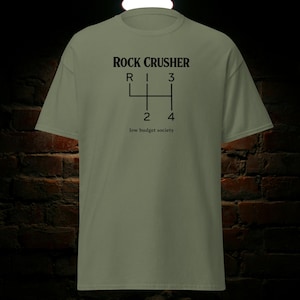 Puede incluir: Camiseta verde oliva con las palabras "ROCK CRUSHER" sobre un gráfico de cambio de marchas negro. Debajo del gráfico está el texto "low budget society". La camiseta está hecha de un material suave.