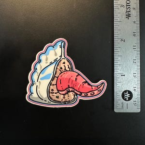 May include: A sticker featuring a cartoon illustration of a creature with a red tongue, a beige body, and a white and blue head. The sticker is approximately 2 inches in length.