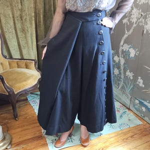 May include: Black culottes with a high waist and button detailing. The pants are paired with a sheer, embroidered blouse. The outfit is completed with brown heels. The background features a vintage-style floral wallpaper and a gold chair.
