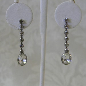 May include: A pair of silver dangle earrings with a row of clear rhinestones and a larger clear oval-shaped rhinestone at the bottom.