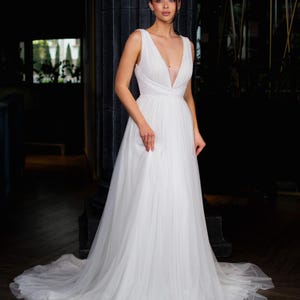 May include: A white wedding dress with a plunging neckline and a flowing skirt. The dress features delicate detailing on the bodice and a long train. The bride is standing in a dark room with a pillar behind her.
