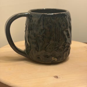May include: A dark, textured ceramic mug with a large handle. The mug has a glossy, mottled finish in shades of black and green. It sits on a light-colored wooden surface.