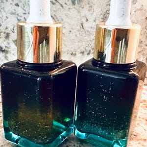 Puede incluir: Dos botellas cuadradas de vidrio con tapas doradas y cuentagotas blancos. Las botellas están llenas de un líquido azul oscuro que se desvanece a turquesa en la base. Las botellas están una al lado de la otra, mostrando un producto para el cuidado de la piel.