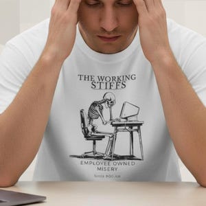 May include: White t-shirt with a graphic of a skeleton at a desk, the text reads "THE WORKING STIFFS EMPLOYEE OWNED MISERY Since 9:00 AM". A person is wearing the shirt and has their hands on their head. The text "Clocked in. Checked out." is at the bottom.