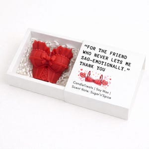 May include: A red soy wax candle shaped like a lingerie top, presented in a white box with decorative paper. The box lid features text that reads "FOR THE FRIEND WHO NEVER LETS ME SAG-EMOTIONALLY. THANK YOU."