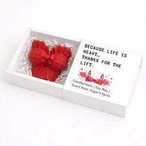 May include: A white box with a red bra-shaped candle inside, nestled on white paper. The box lid has text that reads "BECAUSE LIFE IS HEAVY... THANKS FOR THE LIFT." and a graphic of a bra and hearts. The scent is Sugar'n'Spice.