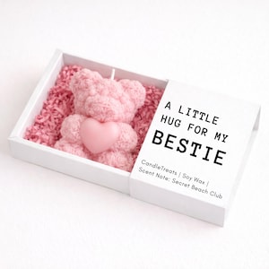 May include: A pink teddy bear candle with a heart, nestled in pink paper, inside a white box. The box lid reads "A LITTLE HUG FOR MY BESTIE". The candle is a decorative item.