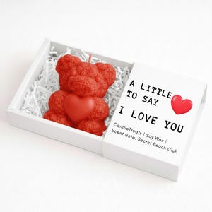 May include: A red teddy bear candle with a heart, presented in a white box with shredded paper. The box lid reads "A LITTLE TO SAY I LOVE YOU" with a red heart graphic. The candle is made of soy wax.