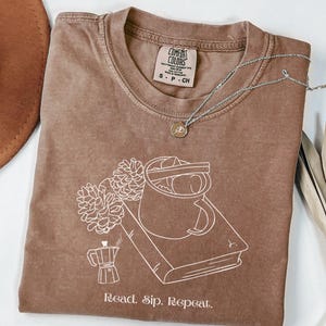 以下が含まれることがあります： コーヒーカップ、本、花が描かれた白いグラフィックの茶色のComfort Colors Tシャツ。「Read. Sip. Repeat.」の文字が下にプリントされています。ペンダント付きのシルバーネックレスがシャツの上にあります。