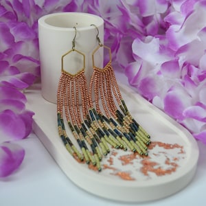 May include: Copper-coloured beaded earrings with a hexagonal top. The earrings feature a fringe of beads in shades of copper, navy, and olive green. They are displayed on a white tray with a copper design, with purple flowers in the background.