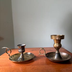 Puede incluir: Dos candelabros de metal con bases circulares y asas laterales. El candelabro de la izquierda es más corto con un acabado pulido, mientras que el de la derecha es más alto con un tallo de tono latón y una base circular más ancha.