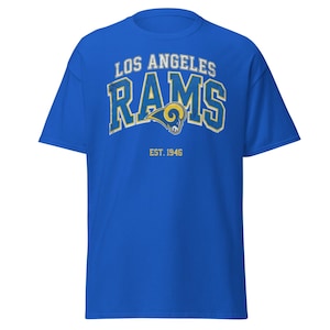 Puede incluir: Una camiseta azul real con el texto "LOS ANGELES RAMS" en blanco y amarillo, un logotipo de carnero y "EST. 1946". La camiseta es de estilo cuello redondo clásico.