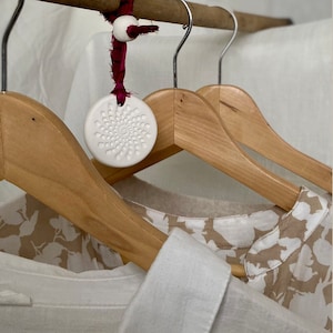 May include: Two wooden hangers with a white ceramic disc hanging from a red ribbon. The disc has a circular pattern of small holes. The hangers are hanging against a white background.