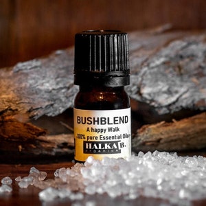 May include: An amber glass bottle of "BUSHBLEND" essential oil with a black cap. The label reads "A happy Walk - 100% pure Essential Oils" and "HALKA B. ORGANICS". The bottle is on a wooden surface with white crystals.