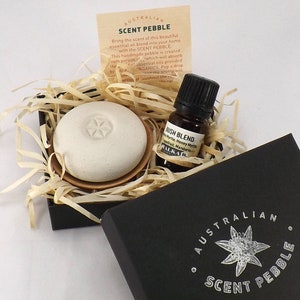 May include: A black box with a white logo that says "Australian Scent Pebble" and a white star with a green centre. The box is open and contains a small white ceramic pebble with a carved design and a small brown glass bottle with a black cap. The bottle is labelled "Bush Blend" and contains essential oils.