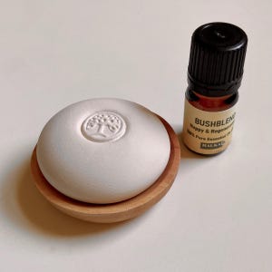 Op de afbeelding: Een witte, ronde diffuser voor etherische olie met een boomontwerp, rustend op een klein houten schaaltje. Daarnaast staat een kleine bruine glazen fles etherische olie met de tekst "Bushblend". De fles heeft een zwarte dop.