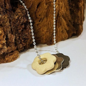 May include: A silver chain necklace with three flower-shaped pendants in beige, silver, and bronze. The pendants are stacked, and the necklace hangs against a textured brown background.
