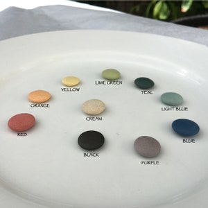 Könnte beinhalten: Ein weißer Teller zeigt elf kleine, ovale Steine in verschiedenen Farben. Die Steine sind mit ihren jeweiligen Farben beschriftet: Rot, Orange, Gelb, Creme, Limettengrün, Türkis, Hellblau, Blau, Schwarz und Lila. Die Steine sind kreisförmig angeordnet.