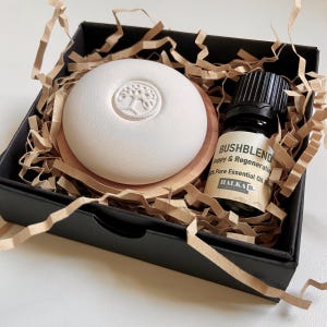 Op de afbeelding: Een geschenkset in een zwarte doos met versnipperd papier. Het bevat een witte, ronde diffuser met een boomontwerp, een houten onderzetter en een kleine fles etherische olie met het label "Bushblend". De oliefles heeft een zwarte dop en een wit etiket.