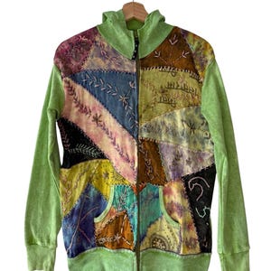 Op de afbeelding: Een zip-up hoodie met een patchwork design. Het patchwork heeft verschillende kleuren, waaronder blauw, geel, roze en bruin, met geborduurde details. De mouwen en de capuchon zijn effen lichtgroen. De hoodie heeft een voorzak.