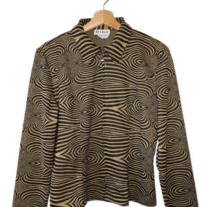 Pode incluir: Casaco bege e preto com zíper e um padrão ondulado semelhante a zebra. O casaco tem mangas compridas e gola. A marca "Dino" é visível na etiqueta. O casaco está pendurado num cabide de madeira.