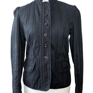 May include: A black jacket with a high collar and vertical striped texture. The jacket has a button-up front, two pockets, and long sleeves. The jacket is displayed on a white mannequin.